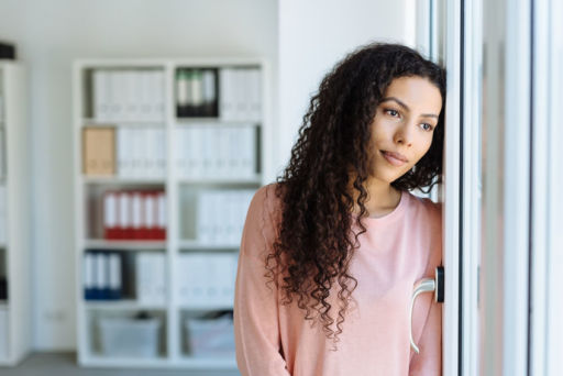 Young woman staring longingly through a window with a sad faraway expression as she leans against a wall in an office